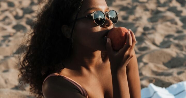 A young woman relaxes on the beach, savoring a peach under the bright sun.