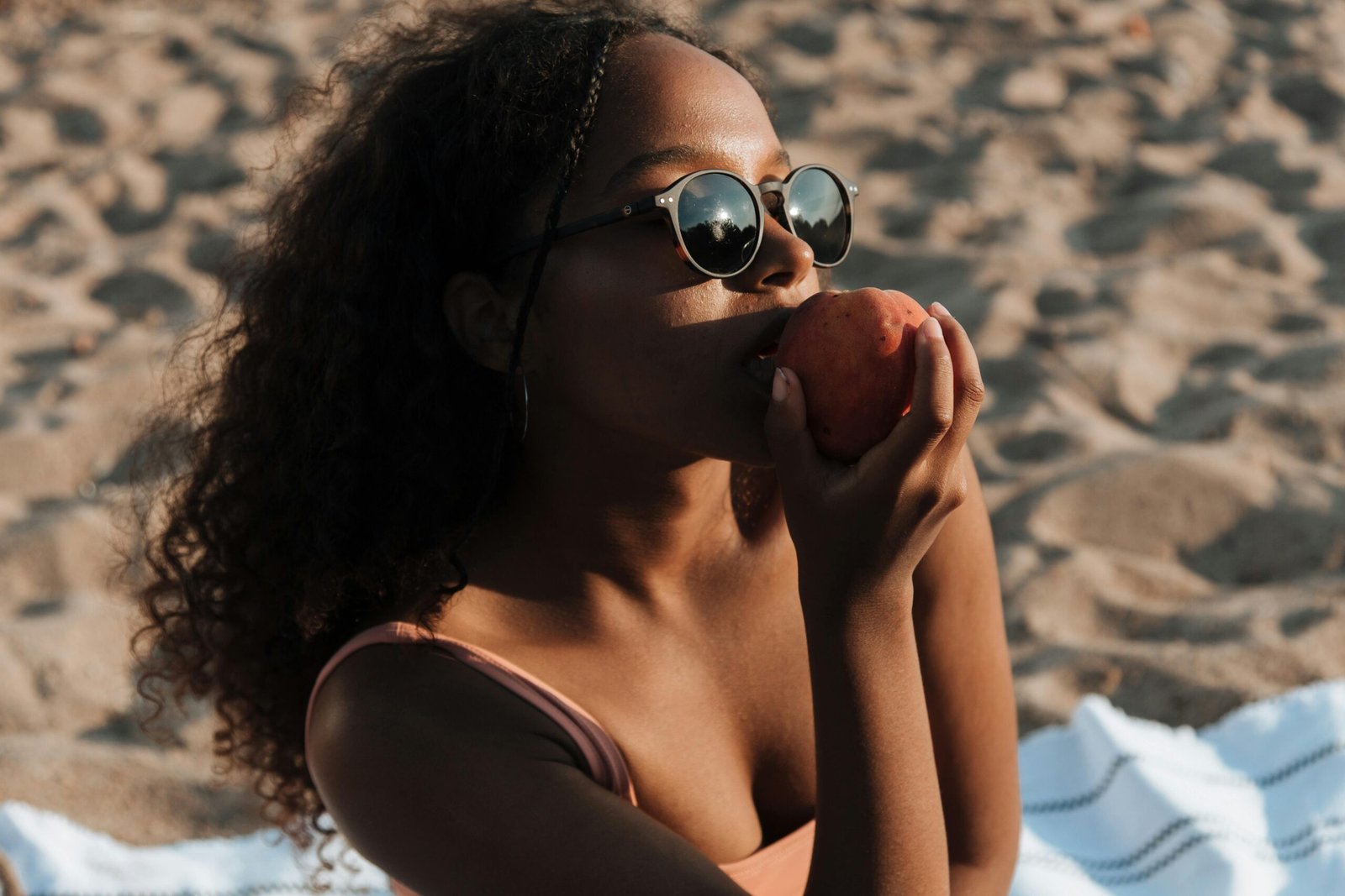 A young woman relaxes on the beach, savoring a peach under the bright sun.