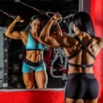Strong female bodybuilder doing cable workout in a gym with mirror reflection.