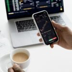 A person trades stocks on a smartphone with a laptop, coffee cup, calculator, and documents in view.